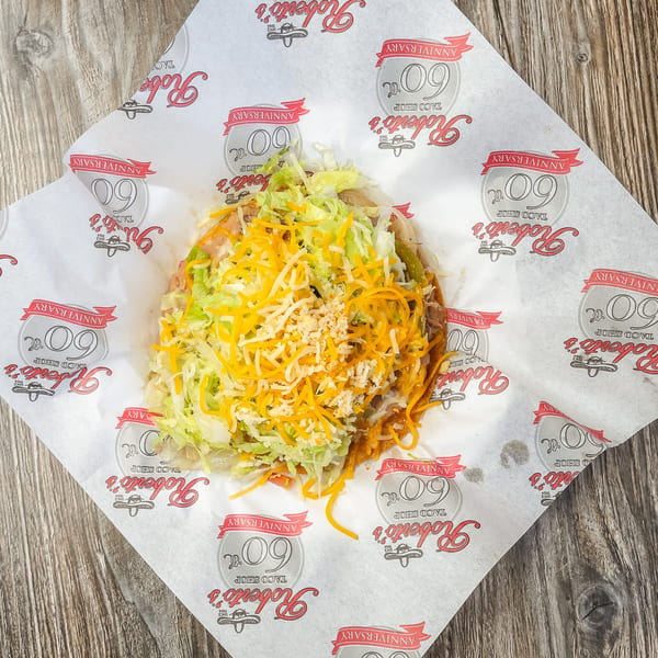 A Roberto's Taco Shop tostada with shredded lettuce and cheese on branded paper.